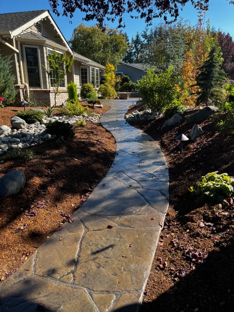 A winding stone path leads through a lush garden, bordered by colorful foliage and sunlit greenery near a charming home.