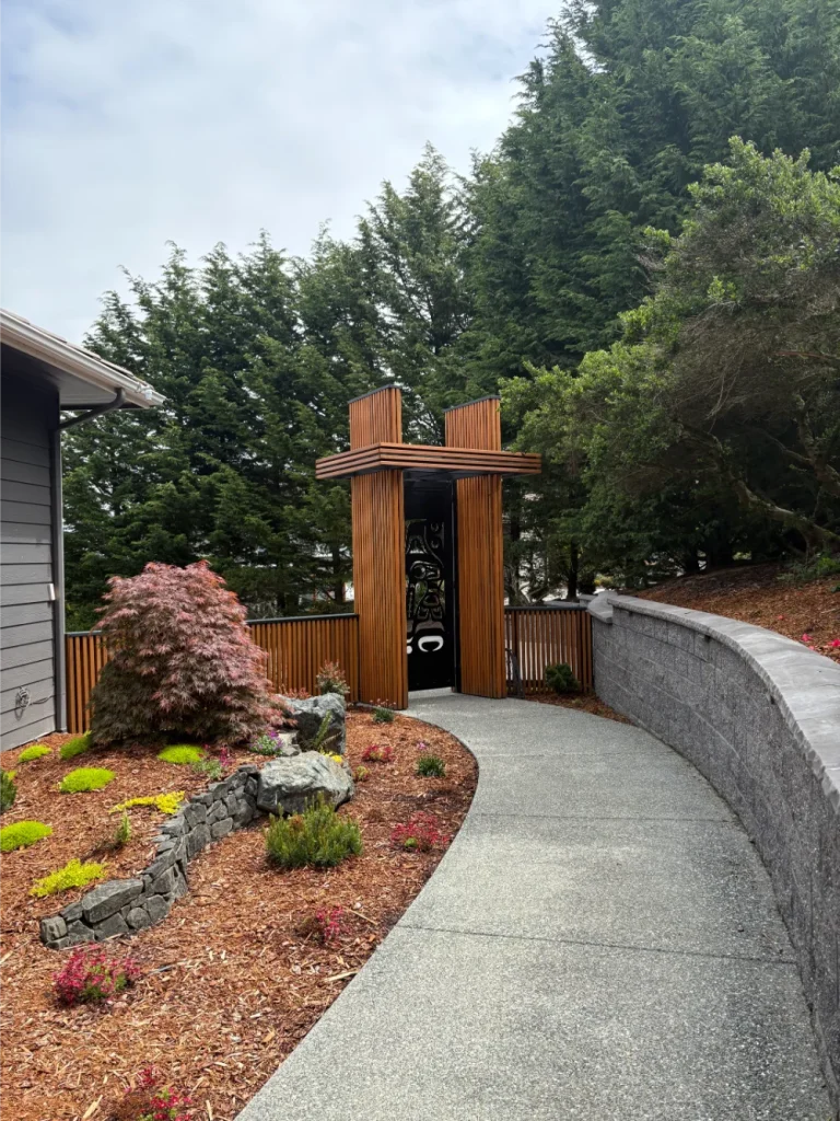 A curved concrete path leads to a wooden gate with a cross design, surrounded by landscaped plants and trees.